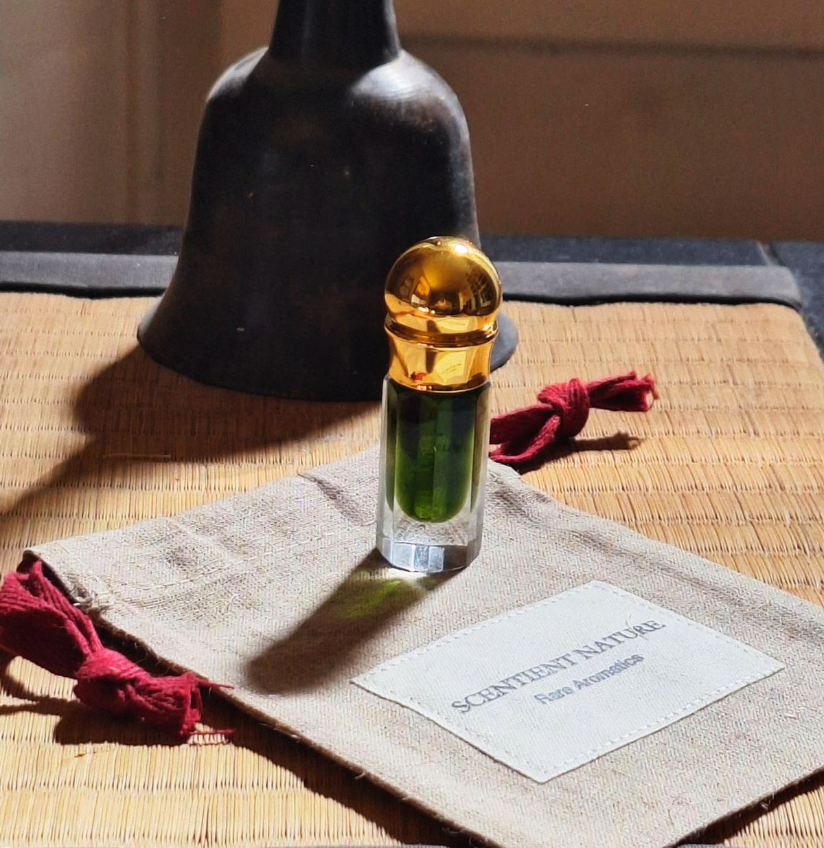 A small glass perfume bottle with a shiny gold cap and emerald green liquid, inula graveolens essential oil, resting on a linen fabric pouch with burgundy tassels. A dark metal bell sits behind it on a woven mat, with natural light casting shadows.